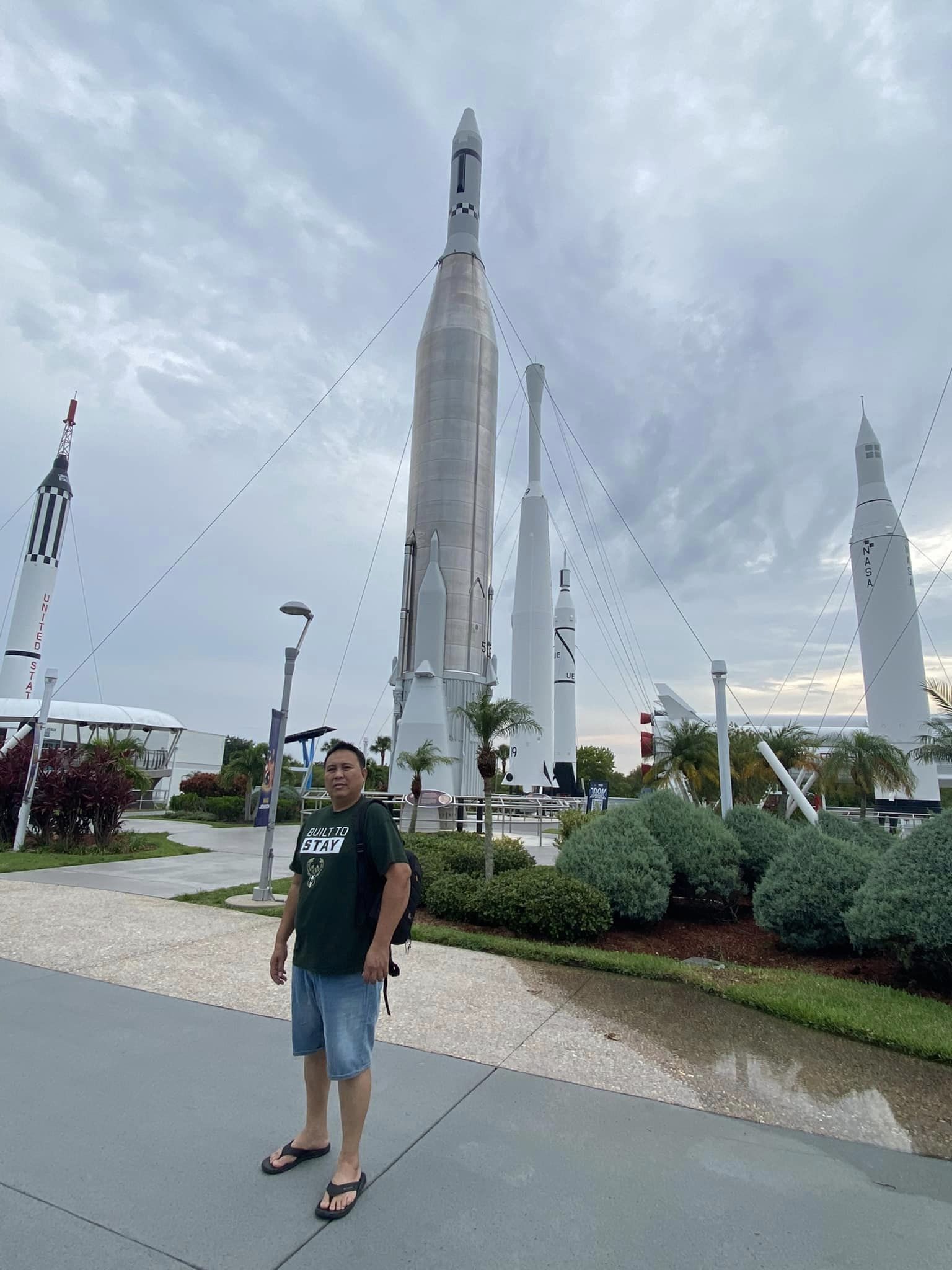 Explorian memberwearing a green shirt and shorts, posing in front of space rockets displayed at an outdoor exhibit.