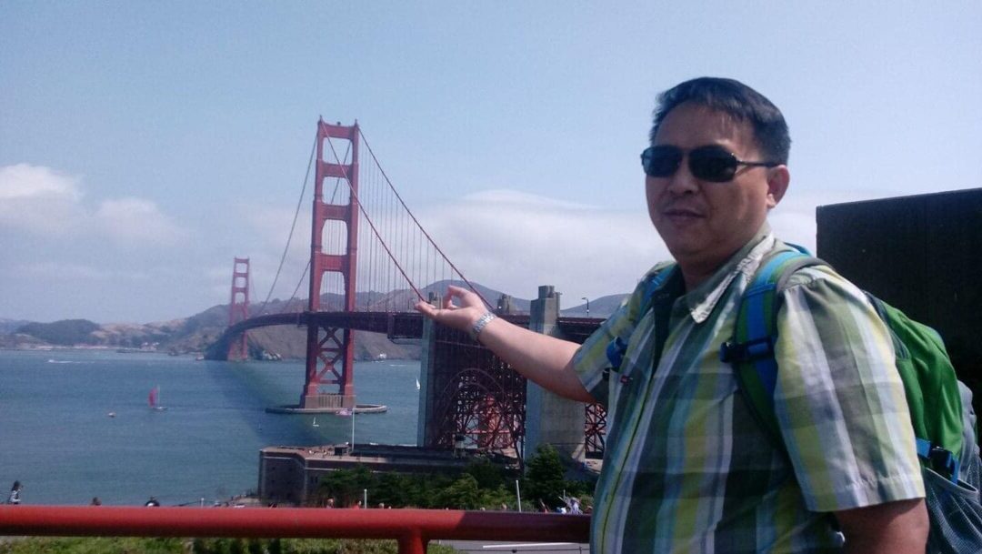 "Exporian member in a checkered shirt and sunglasses, gesturing towards the iconic Golden Gate Bridge in San Francisco."