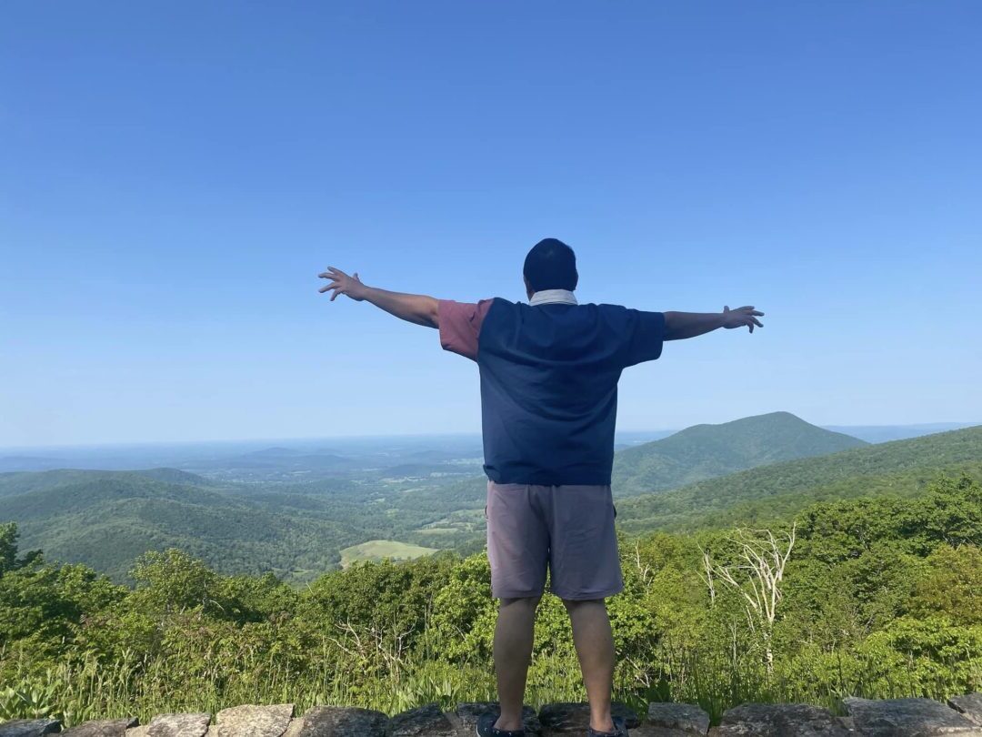 Owner of Explorian standing with arms wide open at the summit of Shenandoah Mountain, West Virginia, in Summer 2023, with a panoramic view of the lush green landscape under a clear blue sky.